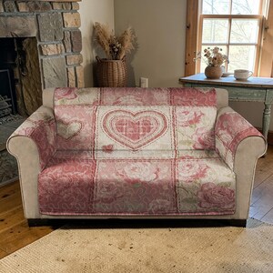 Peut inclure: Une housse de canapé florale rose et blanche avec un motif patchwork. La housse présente de grands appliqués en forme de cœur et des motifs de roses. La housse est placée sur un canapé beige dans un cadre de salon confortable.