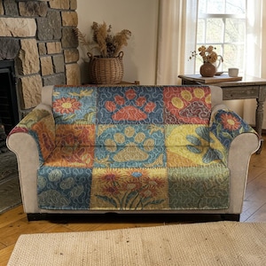 May include: A multi-coloured patchwork sofa cover with floral and paw print patterns. The cover is draped over a beige sofa, and features squares of blue, yellow, orange, and green. The sofa is in a room with a stone fireplace and wooden floor.