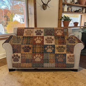 Puede incluir: Un sofá beige con una funda estampada con huellas de patas. La funda presenta un diseño de patchwork con cuadrados de varios colores, incluyendo marrón, verde y azul, cada uno mostrando una huella de pata. El sofá está colocado sobre una alfombra tejida.