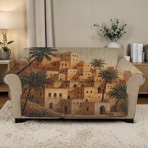 May include: A beige loveseat with a decorative cover featuring a detailed illustration of a Middle Eastern village with palm trees. The cover has a quilted pattern and a warm color palette of browns and tans.