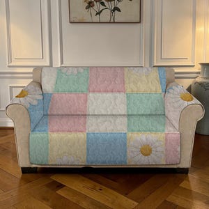 May include: A beige sofa with a colourful patchwork cover. The cover features pastel squares in pink, blue, yellow, and green, with a daisy design on the armrests. The sofa is in a room with a painting and a vase.