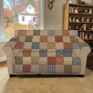 May include: A beige sofa with a patchwork quilt cover featuring squares of various patterns and colors, including red, blue, green, and beige. The sofa is in front of a window with a view of a house and trees.