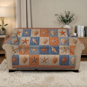 May include: A beige sofa covered with a quilted throw featuring a repeating pattern of seashells and starfish in orange, blue, and beige squares. The throw has a decorative border and is draped over the back and arms of the sofa.