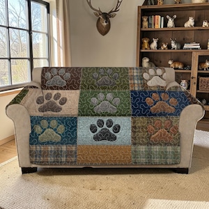 May include: A patchwork sofa cover featuring a repeating paw print design. The cover is made of squares in various colors, including brown, green, and blue, with a paw print in the center of each square. The sofa itself is a neutral beige color.