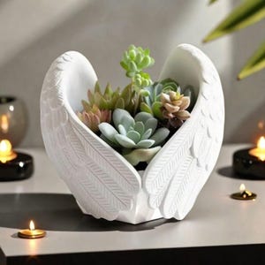 May include: A white, wing-shaped planter filled with various succulents. The planter has detailed feather textures and holds a variety of green, pink, and brown succulents. The arrangement sits on a gray surface, with candles in the background.