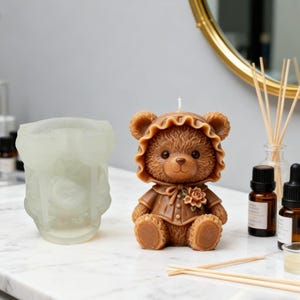 May include: A brown teddy bear candle with a bonnet and a floral accent. The candle is sitting next to a clear mold. The bear is wearing a brown outfit. The candle is on a white surface.
