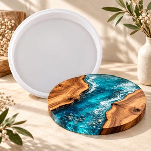 May include: A round white silicone mold positioned behind a circular wooden serving board featuring a blue resin inlay that mimics ocean waves, complete with white foam details and small grey stones embedded within the translucent blue material.