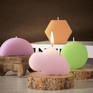 May include: A collection of decorative candles in various shapes and colors. A pink cloud-shaped candle with a lit wick sits on a wooden slice. Other candles include a pink half-circle, an orange hexagon, and a green sphere. The candles are on a neutral background.