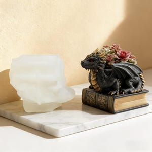 May include: A translucent white silicone mold shaped like a dragon sitting on a book, positioned next to a finished black dragon figurine with gold accents, red eyes, and a succulent plant arrangement resting on top of a dark book with gold detailing.