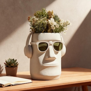 May include: A white, stylized head-shaped planter with sunglasses and headphones, filled with dried flowers and greenery. The planter is on a wooden table, with a small succulent and notebook nearby.
