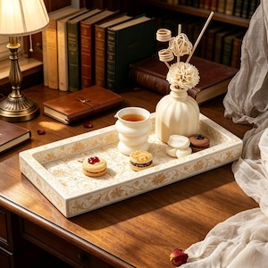 May include: A rectangular ceramic tray with a cream and gold floral pattern holds a white ribbed vase with decorative sticks and a flower, a white cup containing tea, and three small round sandwich cookies with dark and red fillings.