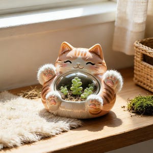 May include: A ceramic cat-shaped planter with a glass globe, holding small succulents. The cat has orange and white stripes, with a smiling face and paw details. The planter sits on a wooden surface.