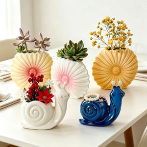 May include: Three round, fluted ceramic vases in yellow, white, and pink, each holding plants. Two snail-shaped ceramic containers are in front: one white holding red berries and flowers, and one glossy blue holding a small pearl-like bracelet.