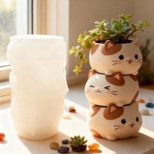 May include: A stack of three white and brown cat-shaped ceramic planters, each with a smiling face. The top planter holds a small succulent plant. A translucent white vase is to the left.