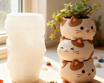 May include: Three stacked, white and brown cat-shaped ceramic plant pots, each with a smiling face. The top pot contains a small succulent plant. A translucent white vase is to the left.