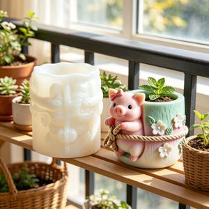 May include: A white silicone mold featuring a detailed embossed floral pattern alongside a finished cylindrical planter with a pink pig figurine, white flowers, and a rope accent, containing a small green plant in soil.