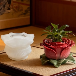 May include: A white frosted glass flower-shaped container sits next to a red ceramic rose-shaped planter containing a small green plant with multiple leaves. The red planter features sculpted petals and green leaves at the base.
