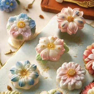May include: Close-up of several flower-shaped pastries. The pastries have intricate petal designs in shades of pink, blue, and white, with yellow centers. Some have green leaf accents. The pastries are arranged on a light blue surface.