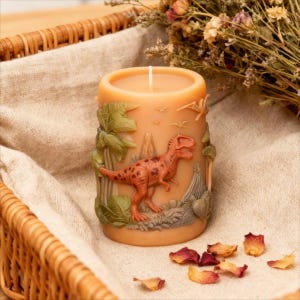 May include: A tan-colored pillar candle with a detailed relief of a Tyrannosaurus Rex, palm trees, and a volcano. The candle has a white wick and is surrounded by dried flowers and rose petals. The candle is approximately 4 inches tall.