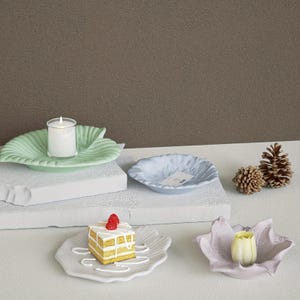 May include: A collection of decorative ceramic dishes and candle holders in various shapes and colors. Includes a leaf-shaped plate with a lit candle, a small cake, and pinecones. The color palette is soft, with pastel greens, blues, and pinks.