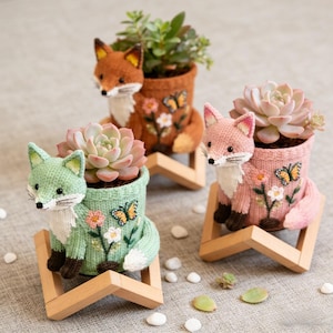 May include: Three knitted fox planters in shades of brown, pink, and green, each holding a succulent plant. The planters are decorated with embroidered flowers and butterflies and sit on small wooden stands. The foxes have white chests and tails.