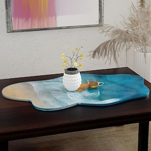 May include: A decorative resin tray with a beach scene design, featuring shades of blue, white, and beige. A small white vase with yellow flowers and a teacup are placed on the tray. The tray is approximately 30 inches long.