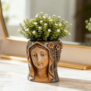 May include: A ceramic planter featuring a serene face with a draped head covering, filled with a green plant and small white flowers. The planter has a brown, tan, and blue color scheme, with floral and grape cluster accents.