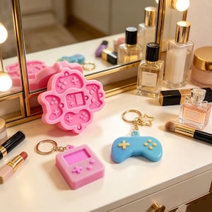 May include: A pink silicone mold featuring four game controller shapes and a central console design, alongside a pink handheld console keychain and a blue game controller keychain with a gold star charm and a metal split ring attachment.