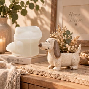 May include: A ceramic dachshund-shaped planter with a speckled cream finish, brown ears, and a brown nose, containing dried flowers. Beside it sits a translucent white silicone mold shaped like a dachshund, resting on a book titled The Art of Home.