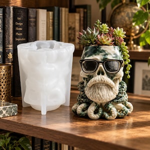 May include: A translucent white silicone mold next to a finished planter shaped like a skull with a beard, wearing black-rimmed glasses, and surrounded by octopus tentacles. The skull planter contains a variety of small green and red succulents.