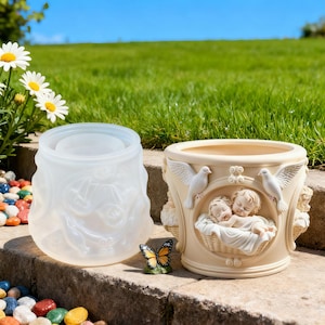 May include: A translucent silicone mold and a finished cream-colored cylindrical planter featuring a relief design of two sleeping infants in a basket, flanked by two white doves, with a small butterfly figurine placed between the two items.