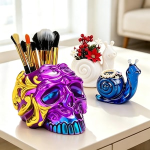 May include: A purple and gold skull-shaped holder containing several makeup brushes, accompanied by a white snail-shaped planter with red berries and a blue snail-shaped trinket dish holding a pearl bracelet and small jewelry items.