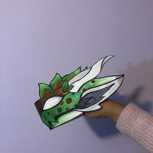 May include: A handmade, colorful dragon mask with green, brown, white, and gray accents. The mask features a detailed design with a textured appearance, and is held up by a person's hand. The mask is made of paper and is suitable for costume parties.