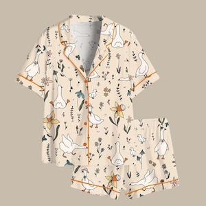 May include: A two-piece pajama set featuring a cream-colored background with a whimsical pattern of white geese, flowers, and foliage. The set includes a short-sleeved button-up shirt and matching shorts, both trimmed with orange piping.