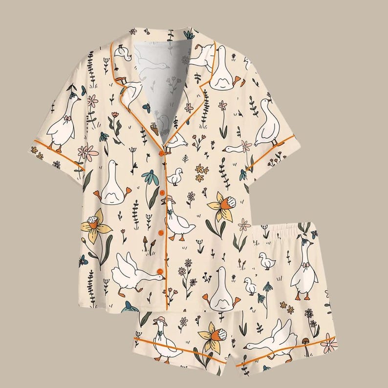 May include: A two-piece pajama set featuring a cream-colored background with a whimsical pattern of white geese, flowers, and foliage. The set includes a short-sleeved button-up shirt and matching shorts, both trimmed with orange piping.