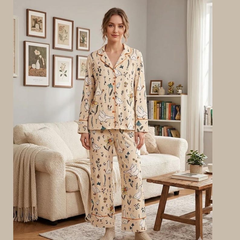 May include: Beige pajama set with a white goose and floral pattern. The long-sleeved top has a collared neckline and button-down front. The pants have a matching print and a relaxed fit. The trim is a contrasting orange color.