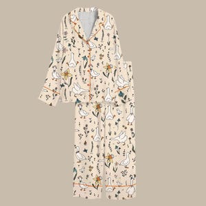 May include: Cream-colored pajama set featuring a whimsical pattern of white geese, flowers, and foliage. The long-sleeved top has orange trim and buttons, paired with matching pants. A comfortable sleepwear set.