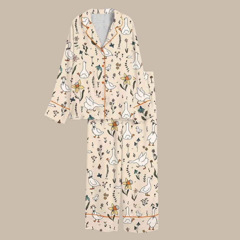 May include: Cream-colored pajama set featuring a whimsical pattern of white geese, flowers, and foliage. The long-sleeved top has orange trim and buttons, paired with matching pants. A comfortable sleepwear set.
