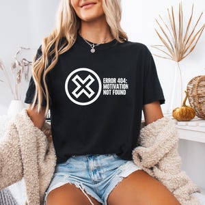 May include: Black t-shirt with the text "ERROR 404: MOTIVATION NOT FOUND" in white. The shirt features a white graphic of an "X" inside a circle. The model is wearing a beige cardigan and blue denim shorts.