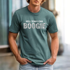 Può includere: T-shirt girocollo color verde acqua con la scritta "HELL YEAH I CAN BOOGIE" in un font glitterato argento. La maglietta è a tinta unita e ha maniche corte.