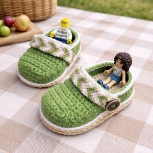 May include: Two green crochet baby shoes with white and tan accents. Each shoe has a strap with a chevron pattern and a button closure. Small toy figures are inside the shoes. The shoes are on a checkered surface.