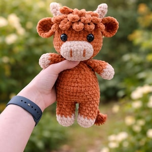 May include: A handmade crochet Highland cow plushie in shades of brown and beige. The plush toy has a fluffy, textured appearance with small horns, ears, and a tail. The cow is held against a blurred green and yellow background.
