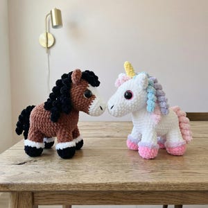 May include: Two crocheted stuffed animals: a brown horse with a black mane and a white-faced head, and a white unicorn with a rainbow mane and pink hooves. Both are sitting on a wooden table.