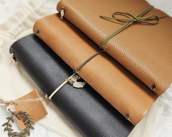 May include: Three leather journals in shades of brown and black are stacked on a white surface. The top journal is a light brown with a green tie closure. The middle journal is a medium brown, and the bottom journal is black. Each journal has a decorative charm.