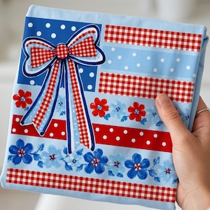 Può includere: Un tessuto azzurro piegato con un design patriottico. Presenta un grande fiocco con motivo a quadri rosso e bianco e nastri blu. Motivi floreali e a pois rossi e blu completano il design.