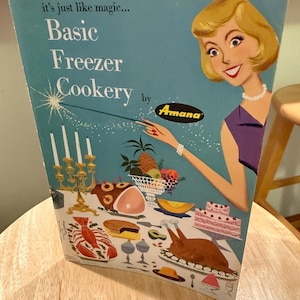 May include: Vintage cookbook cover titled "Basic Freezer Cookery" by Amana. The cover features a cartoon illustration of a woman in a purple dress, pointing a magic wand at a table laden with food. The background is a light blue color.