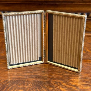 May include: A gold-colored, hinged picture frame, open to reveal two rectangular spaces with corrugated cardboard backing. The frame has a decorative, patterned border. The frame is displayed on a wooden surface.