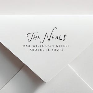 May include: White envelope with black text. The text reads "The Neals" in a cursive font, followed by the address: 263 Willoughby Street, Arden, IL 58216. The envelope is folded at an angle.