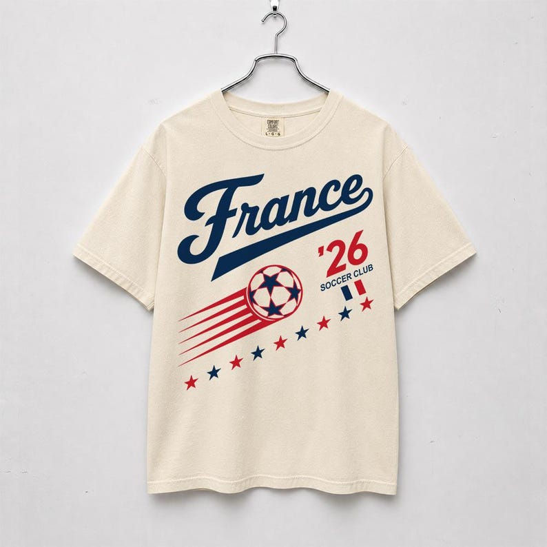 May include: Cream-colored t-shirt with the word "France" in navy blue script. Below, "'26 SOCCER CLUB" is printed, along with a soccer ball and red and blue star accents. The shirt is hanging on a metal hanger.