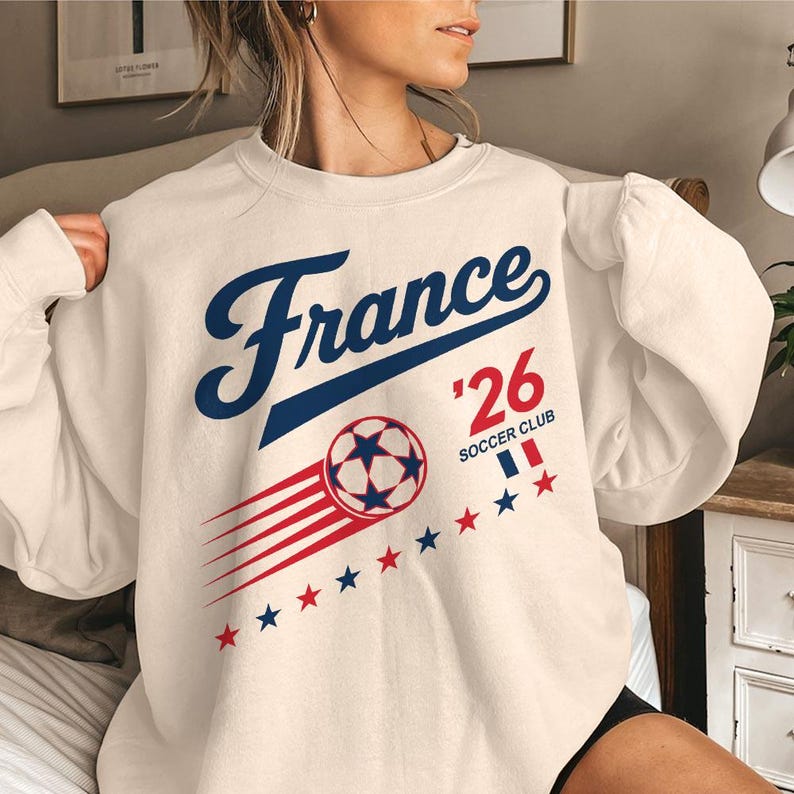 May include: Cream-colored sweatshirt with the word "France" in blue script, "'26" in red, and a soccer ball graphic. Features red and blue star accents and the text "Soccer Club".